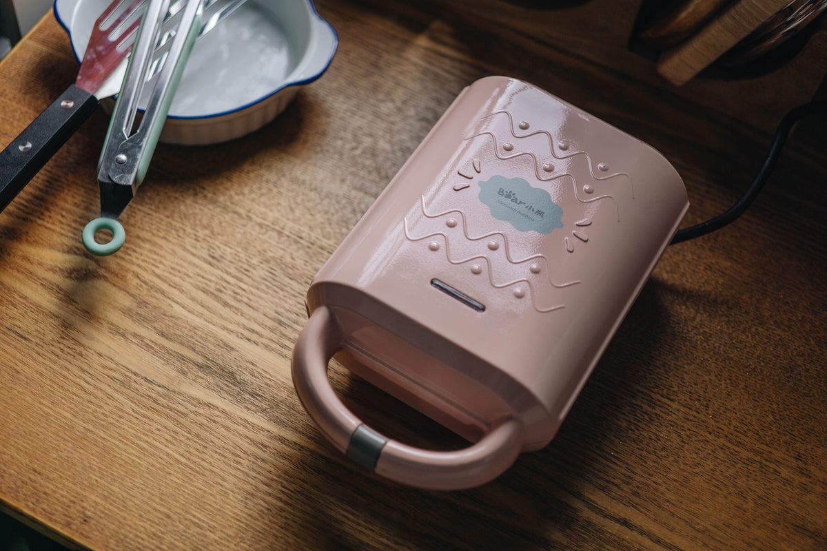 Pink sandwich maker on wooden table with kitchen utensils, perfect for cozy and modern kitchen setting.